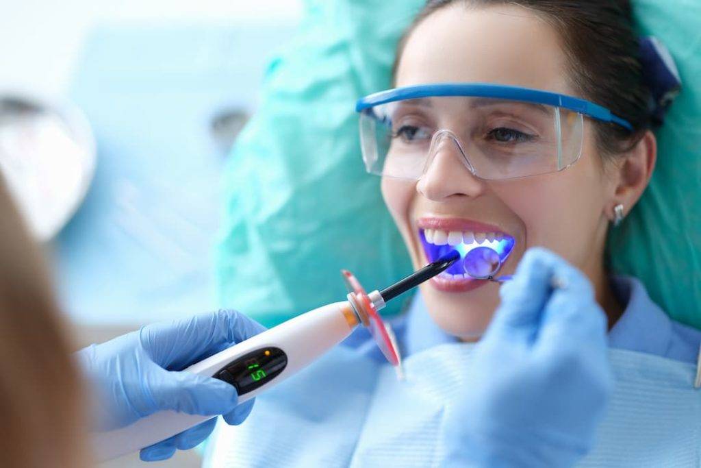 Woman With Uv Lamp In Her Mouth Getting Composite Bonding Applied By A Dentist